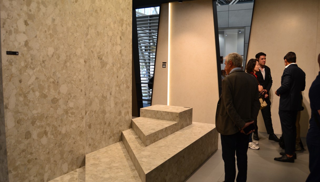 Stand fieristico con pareti e gradini rivestiti in lastre effetto graniglia dai toni sabbia, con visitatori che osservano i materiali