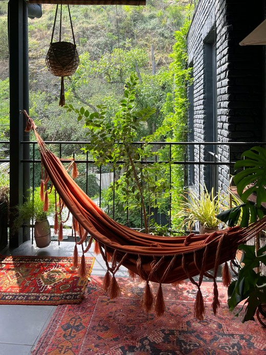 Veranda coperta con amaca in tessuto ruggine e frange, tappeti persiani sovrapposti, cesto sospeso e piante in vaso affacciate sul verde.