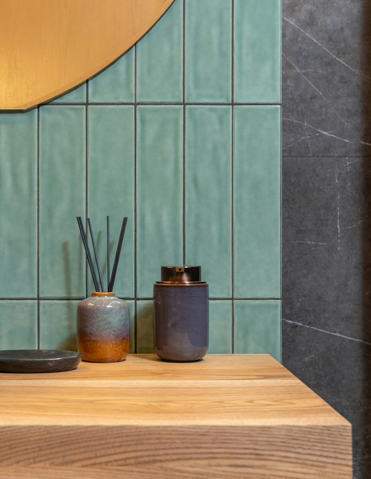 Angolo di bagno con piastrelle rettangolari verde salvia posate in verticale, piano in legno chiaro, ceramiche smaltate e specchio tondo dorato.