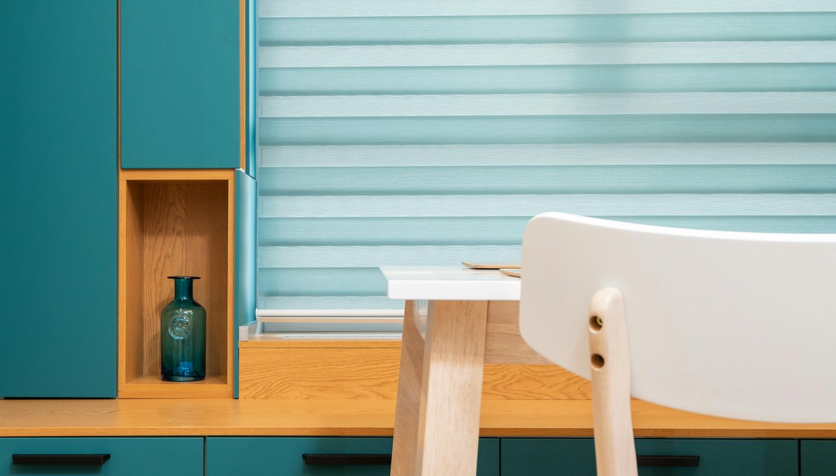 Cucina con ante verde petrolio e maniglie lineari nere, piano e nicchia in legno chiaro con bottiglia in vetro blu, tenda a pacchetto azzurra e sedia bianca