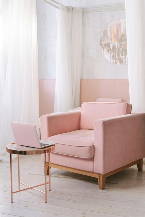 Poltrona in velluto rosa cipria accanto a un tavolino tondo in rame con laptop, tra tende bianche leggere e pareti con boiserie rosa e quadro circolare.