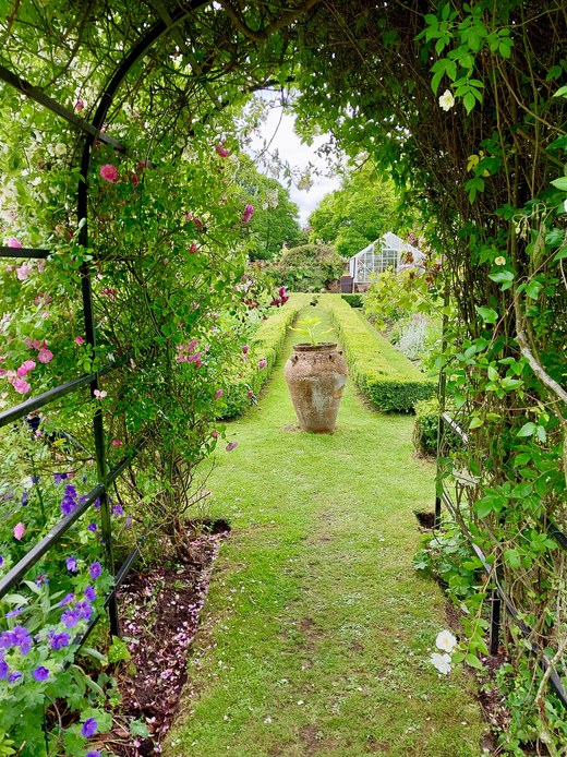 Vialetto erboso visto attraverso un arco di rose rampicanti, con un grande vaso in terracotta al centro, bordure fiorite laterali e una serra bianca in fondo