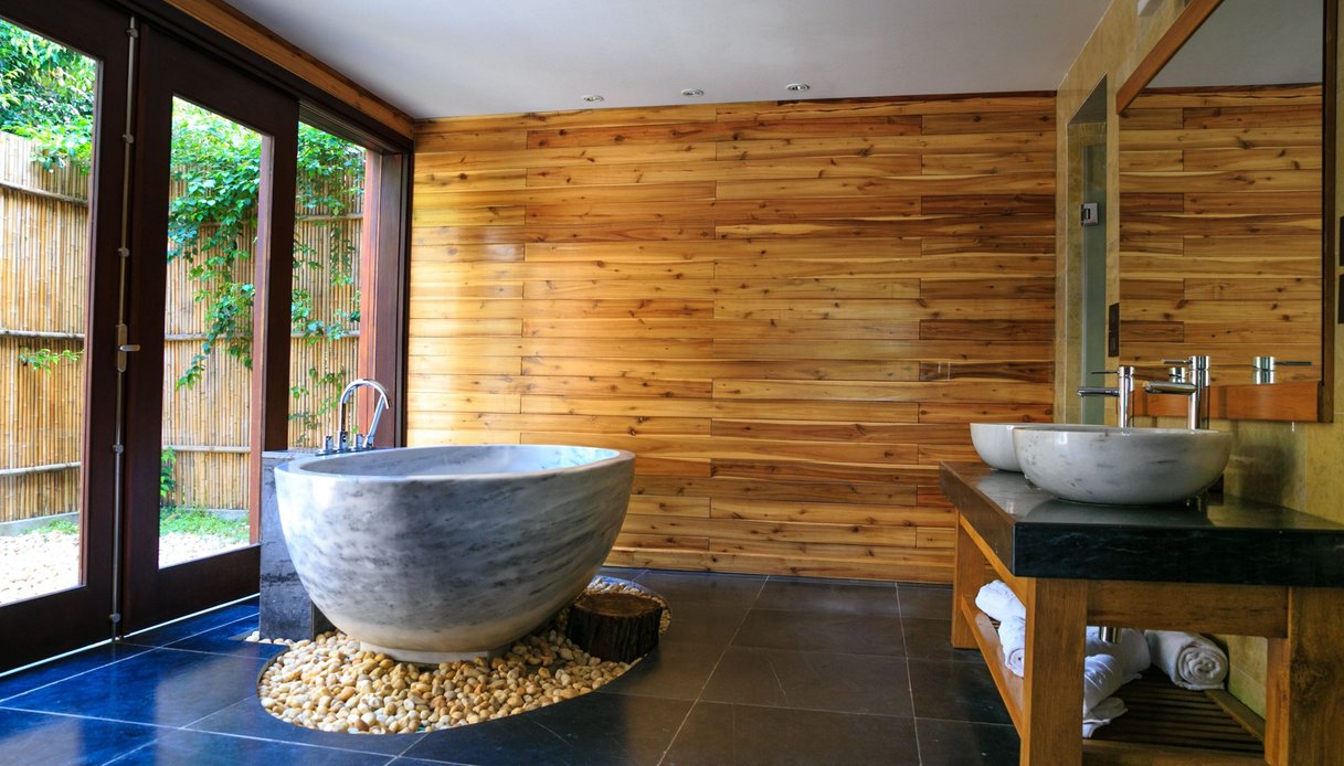 Bagno con parete in doghe di legno, vasca freestanding in pietra grigia su un letto di ciottoli, pavimento in ardesia scura, doppio lavabo d’appoggio e vetrate su un giardino con bambù