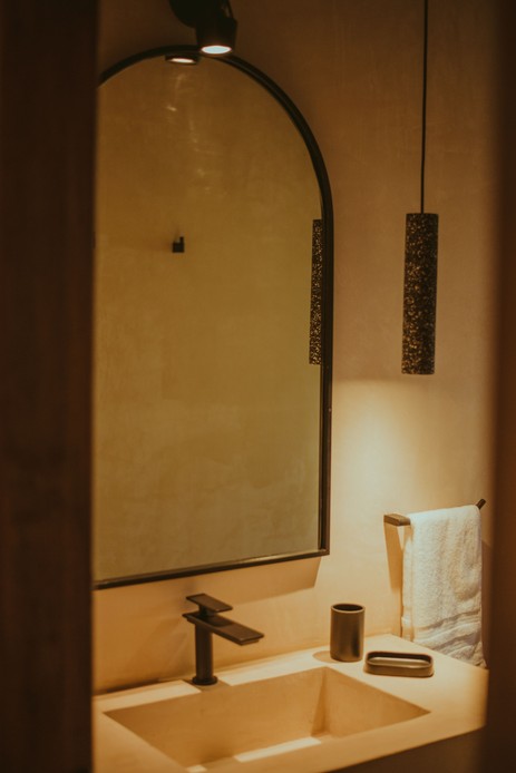 Bagno con lavabo integrato in microcemento color avorio, rubinetto nero, specchio ad arco e lampada a sospensione cilindrica