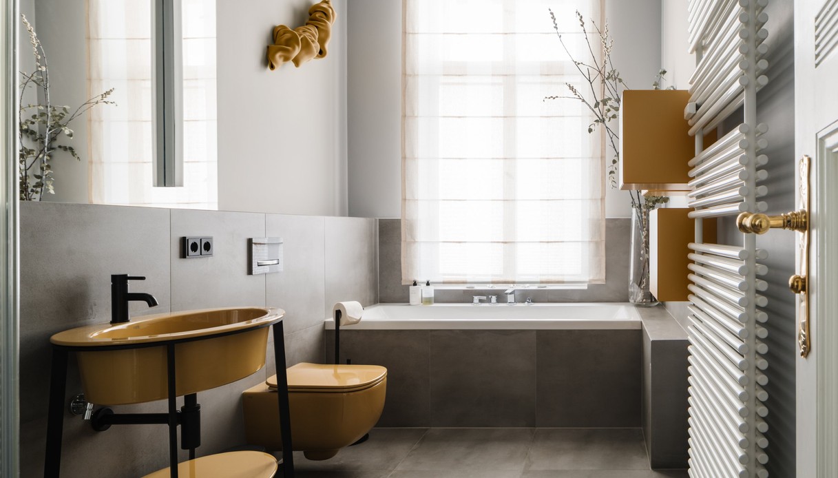 Bagno con lavabo da appoggio e sanitario sospeso color senape, struttura metallica nera, rivestimento in grandi lastre grigie, vasca sotto la finestra con tenda chiara, scultura gialla a parete e termoarredo bianco