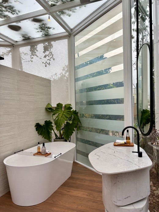 Bagno vetrato con vasca freestanding bianca su parquet, grande pianta di philodendron, lavabo in marmo a colonna con rubinetto nero e specchio ovale, sotto un tetto in vetro con foglie
