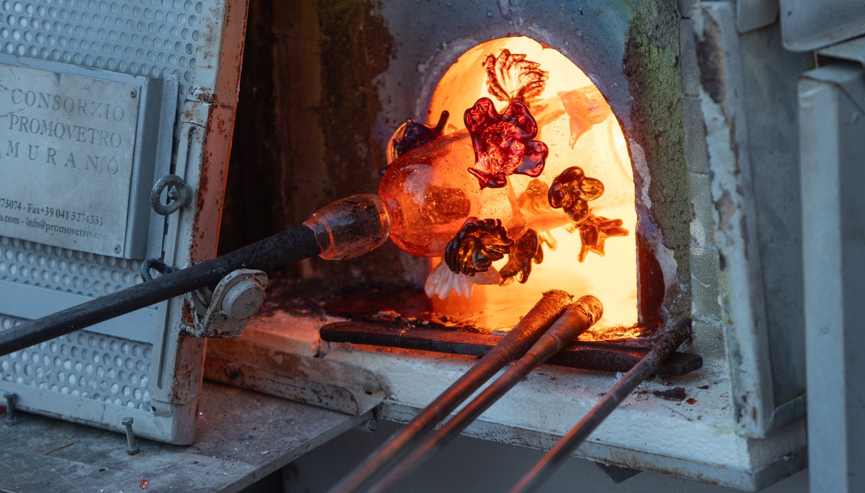Bocca di una fornace aperta con una massa di vetro incandescente arancione infilata sulla canna da soffio, in un laboratorio di Murano.