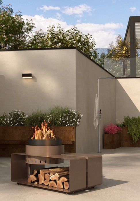 Braciere acceso con legnaia integrata in metallo color bronzo in un patio con muri intonacati, fioriere in acciaio corten e doccia da esterno in acciaio con telo appeso