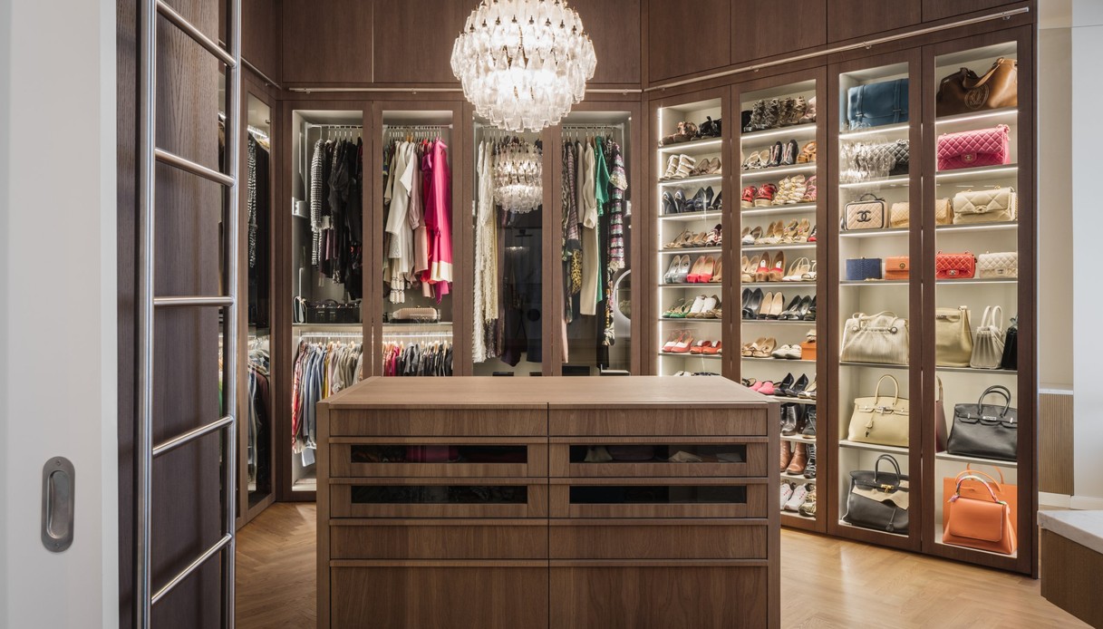 Cabina armadio in legno scuro con ante in vetro illuminate che espongono abiti appesi, scarpe e borse, isola centrale con cassetti e grande lampadario in vetro a sfera, vista attraverso una porta scorrevole