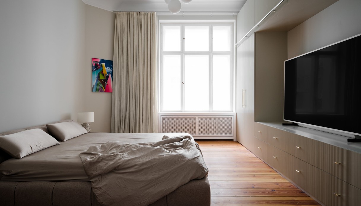 Camera da letto con letto imbottito tortora e lenzuola in tinta, grande finestra con tenda in lino, quadro astratto colorato alla parete, armadio e mobile tv su misura con frontali scanalati e pavimento in tavolato di legno