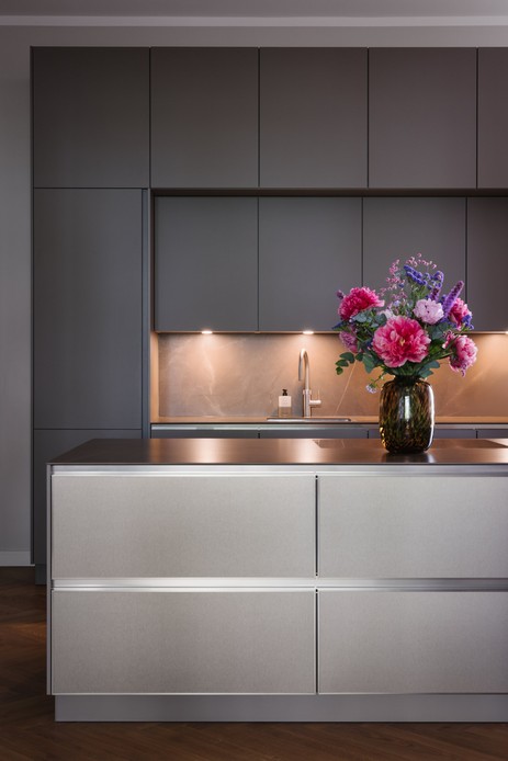 Cucina con colonne e pensili grigi opachi, alzatina in pietra venata illuminata dal basso, rubinetto in acciaio e isola con frontali in acciaio spazzolato, su cui poggia un vaso con peonie rosa