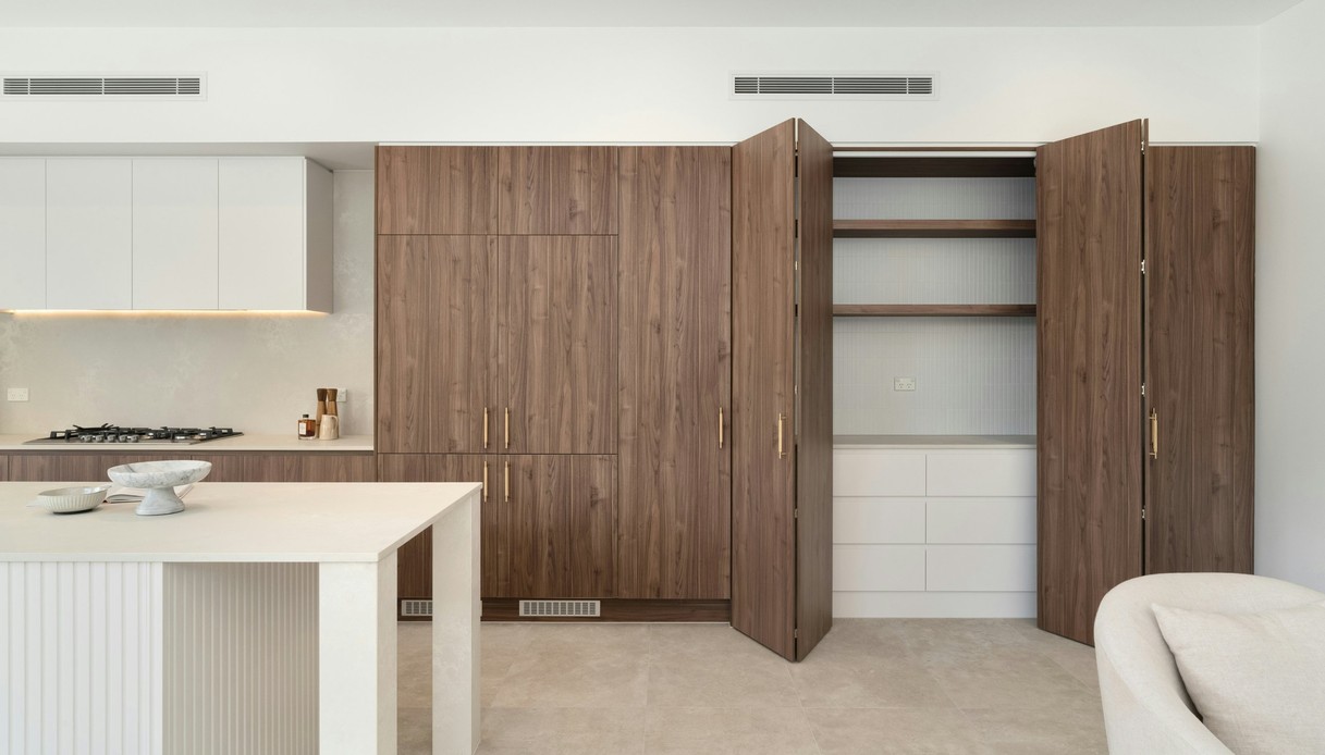 Cucina con ante in legno di noce a tutta altezza che incorniciano un grande frigorifero doppio in acciaio, isola con piano chiaro a cascata e pavimento in legno con tappeto