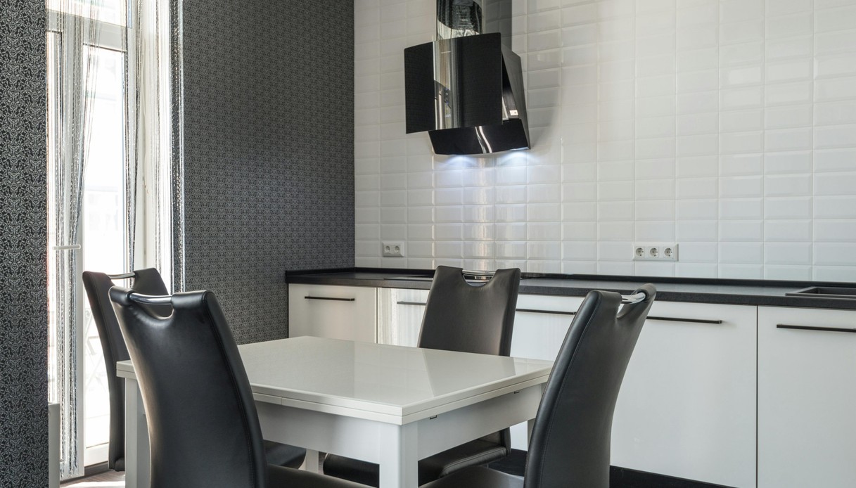 Cucina con rivestimento in piastrelle diamantate bianche, mobili bianchi con piano nero, cappa nera inclinata, tavolo bianco con sedie in pelle nera e carta da parati scura a motivi