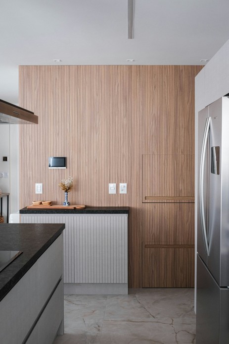 Parete di cucina rivestita in doghe di legno chiaro con ante a filo quasi invisibili, mobile basso cannettato bianco con piano in granito scuro e frigorifero in acciaio di lato