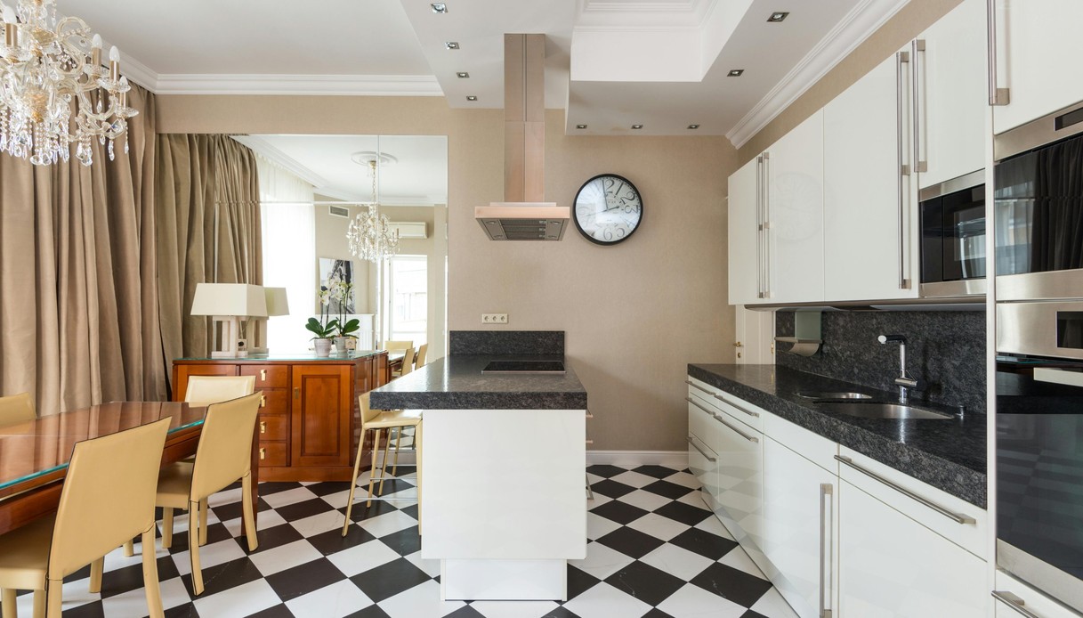 Cucina con pavimento a scacchiera bianco e nero posato in diagonale, isola bianca con piano in granito scuro, mobili laccati bianchi e sala da pranzo con lampadario di cristallo