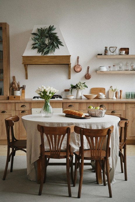 Cucina in legno con tavolo rotondo coperto da una tovaglia lunga in lino naturale, sedie in legno curvato, tulipani bianchi in un vaso di vetro, pane e cestino di pere