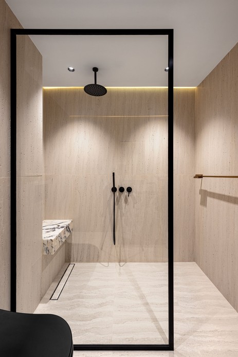 Doccia walk-in rivestita in travertino con parete in vetro dal profilo nero, soffione a pioggia a soffitto, rubinetteria nera, panca in marmo venato e luce LED nascosta lungo il soffitto