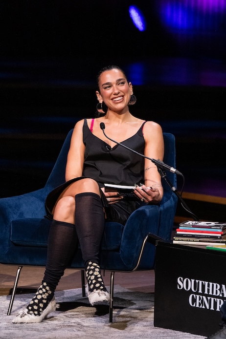 Dua Lipa al London Literature Festival 2026 con completo nero Prada, calzini al ginocchio decorati da margherite e scarpe con piume bianche.