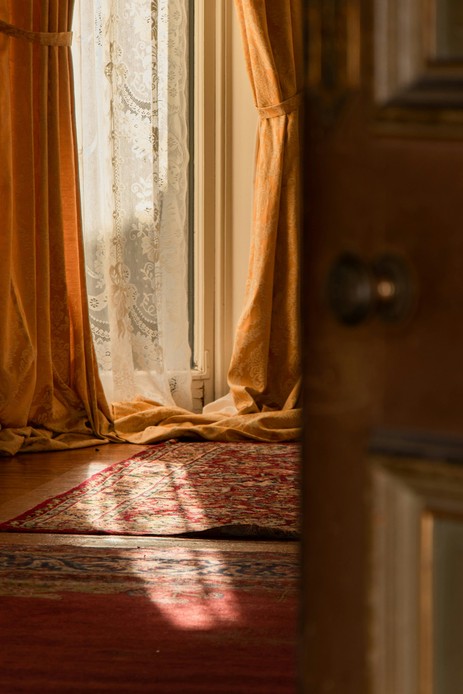 Porta finestra con tende damascate color ocra raccolte da fermatende e tenda di pizzo bianco, luce calda sul parquet e su due tappeti persiani, con una porta in legno in primo piano