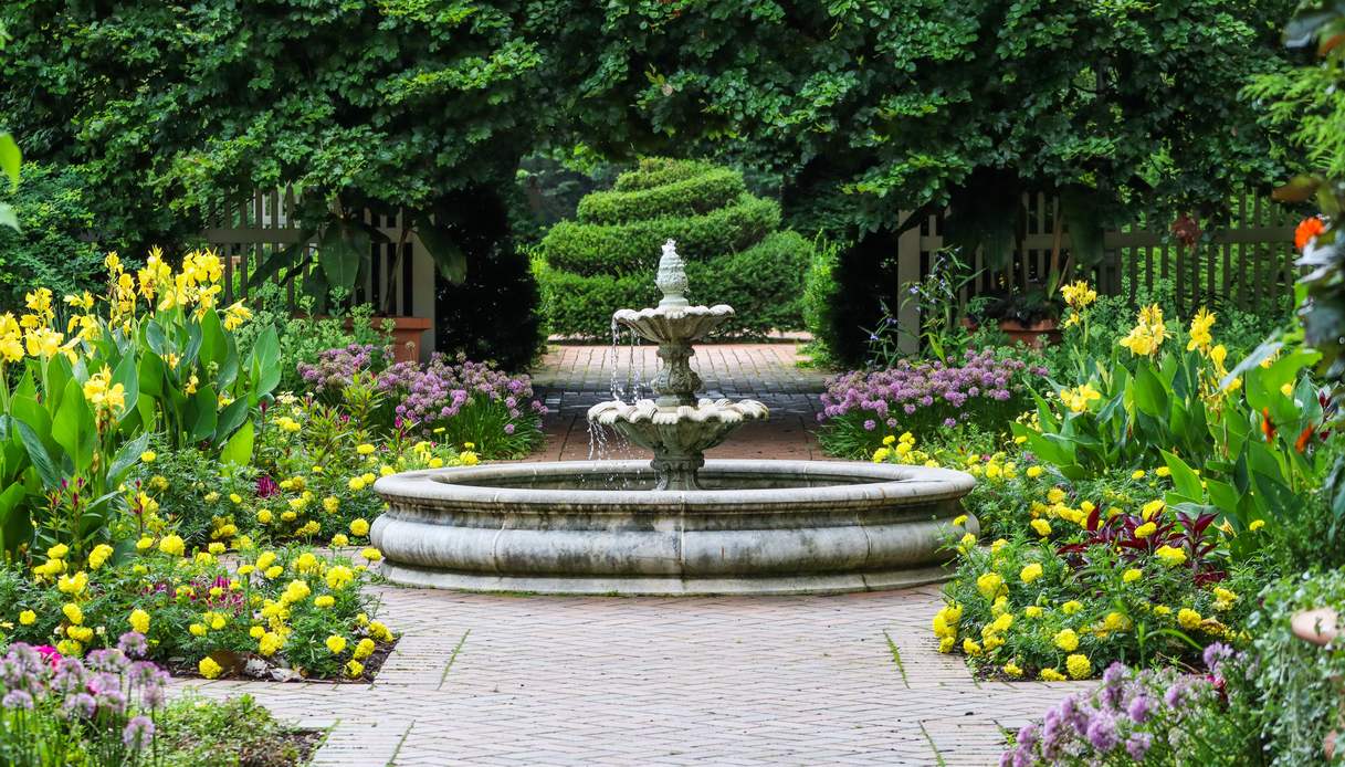 Fontana in pietra a due vasche al centro di un vialetto in mattoni, tra aiuole con fiori gialli e lilla e siepi potate