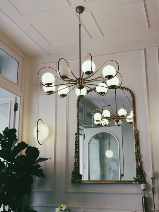 Lampadario in ottone con bracci curvi e sfere in vetro opalino, applique abbinata e grande specchio con cornice dorata intagliata su una parete bianca con boiserie