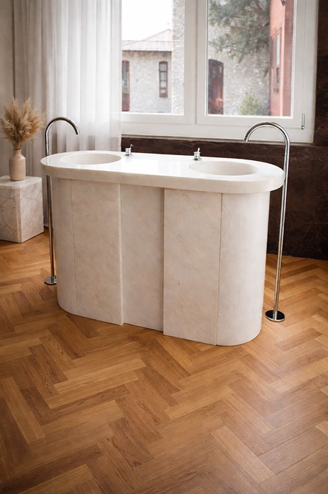 Lavabo doppio freestanding in pietra chiara dalle forme arrotondate, con rubinetti a colonna su parquet in legno posato a spina di pesce