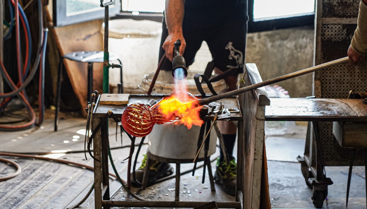 Lavorazione del vetro in fornace: una fiamma riscalda un pezzo ambrato montato su un supporto metallico, tra attrezzi e banchi da lavoro.