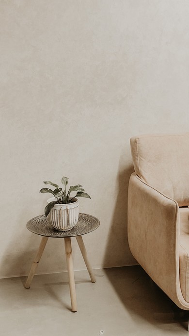 Angolo di soggiorno con parete e pavimento in microcemento beige, tavolino tripode con pianta in vaso e poltrona color sabbia