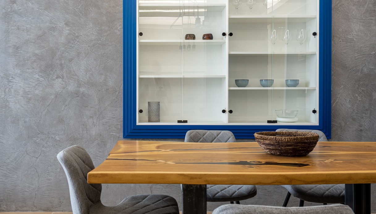 Zona pranzo con parete in microcemento grigio a spatola, vetrina incassata con cornice blu, tavolo in legno con inserti in resina e sedie imbottite grigie