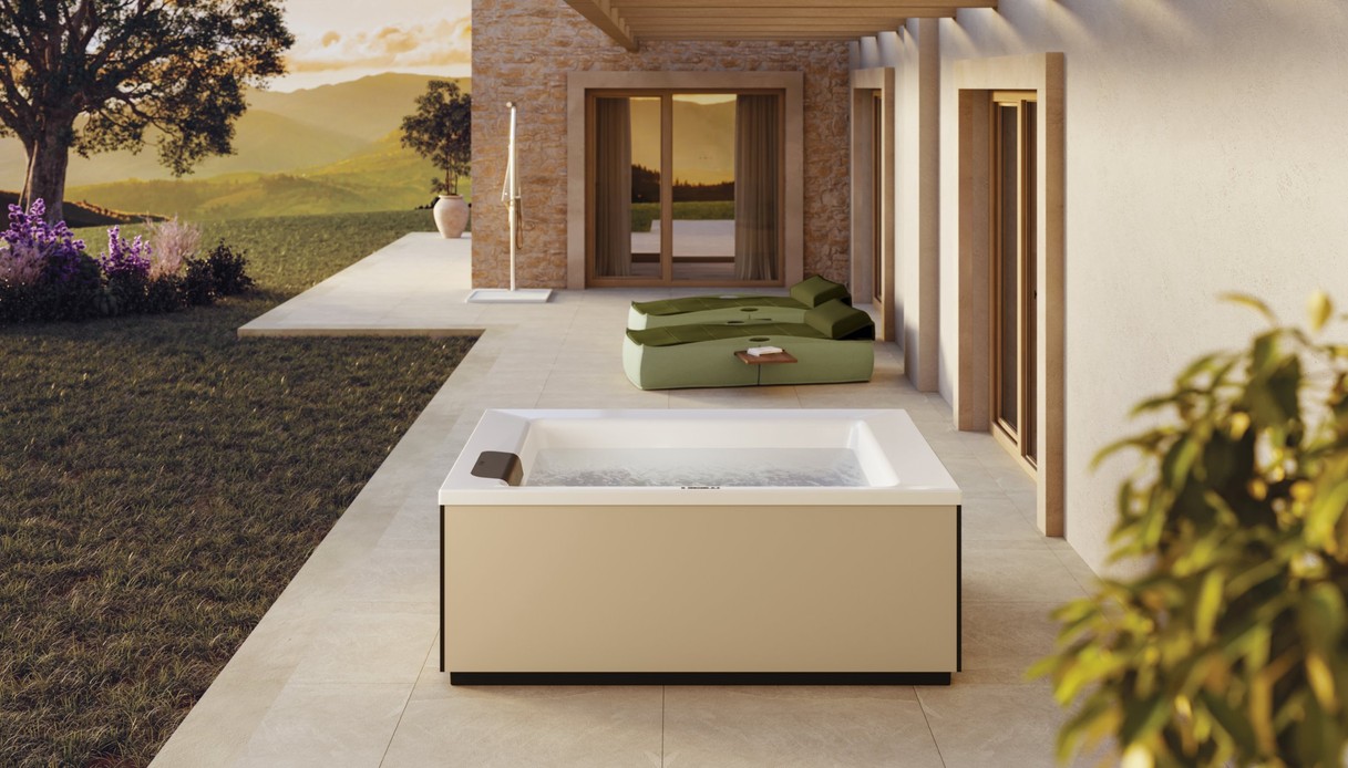 Minipiscina idromassaggio con fianchi rivestiti color sabbia sotto il portico di una casa in pietra, con prendisole verdi, doccia da esterno e colline al tramonto sullo sfondo