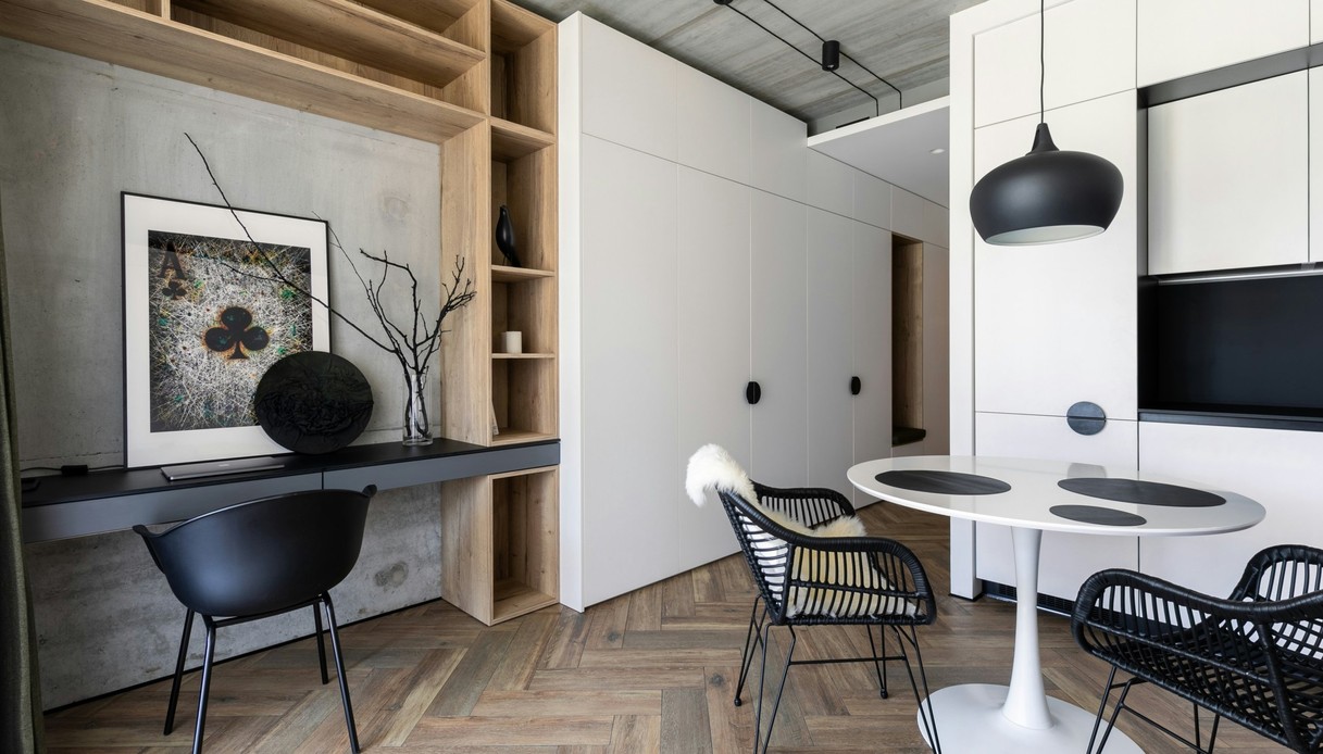 Monolocale con parete in cemento a vista, libreria in legno chiaro, scrivania nera, armadiature bianche, tavolo tondo bianco con sedie in rattan nero e parquet a spina di pesce