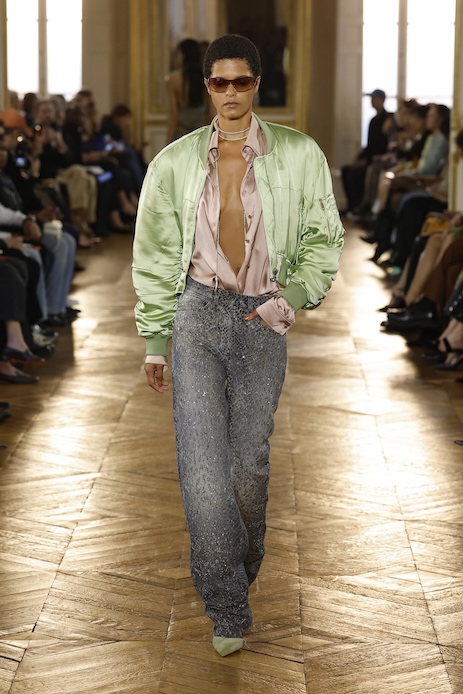 Modella alla sfilata Balmain durante la Paris Fashion Week con jeans luminosi, camicia satinata rosa cipria e bomber verde pastello.