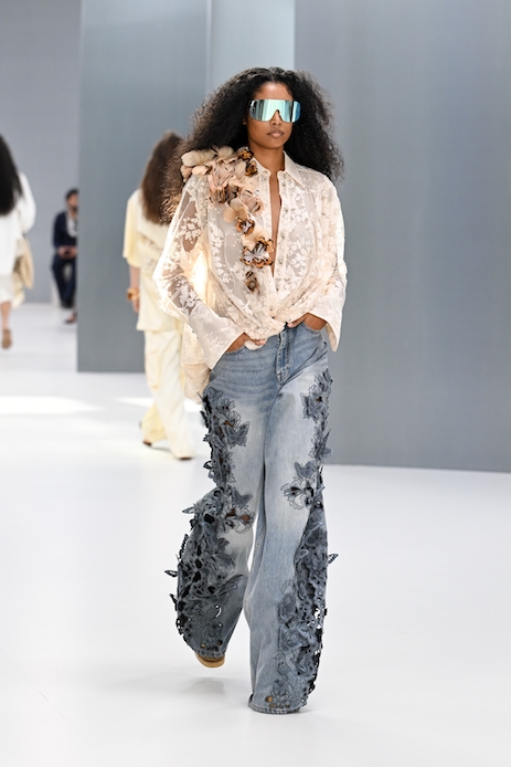 Modella alla sfilata Zimmermann durante la Paris Fashion Week con camicia in pizzo bianco, jeans azzurri con applicazioni floreali e occhiali specchiati.