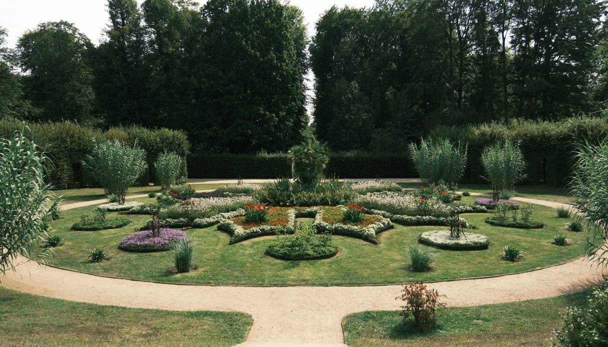 Parterre simmetrico con aiuole fiorite disposte a disegno regolare sul prato, graminacee alte ai lati, vialetti in ghiaia e alte siepi di fondo