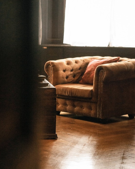 Poltrona Chesterfield in velluto color cammello con borchie e cuscino rosso, accanto a una cassapanca in legno scuro su parquet a spina di pesce, in controluce davanti a una finestra