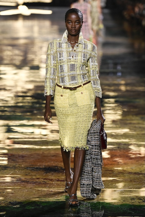 Rachel Ojuromi debutta sulla passerella Chanel a Parigi con un look composto da giacca corta strutturata, gonna midi in tweed giallo, cintura sottile e sandali metallici.