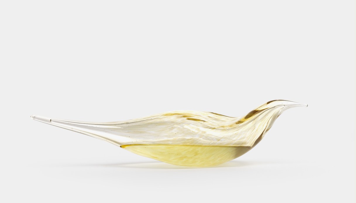 Scultura in vetro dalla forma allungata e affusolata nei toni dell'ambra e del miele, fotografata su fondo chiaro uniforme.