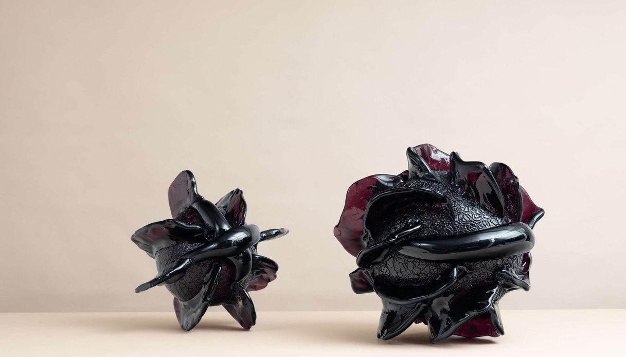 Due sculture in vetro dalle superfici scure e dalle forme organiche accartocciate, con riflessi vinaccia, su un piano chiaro.