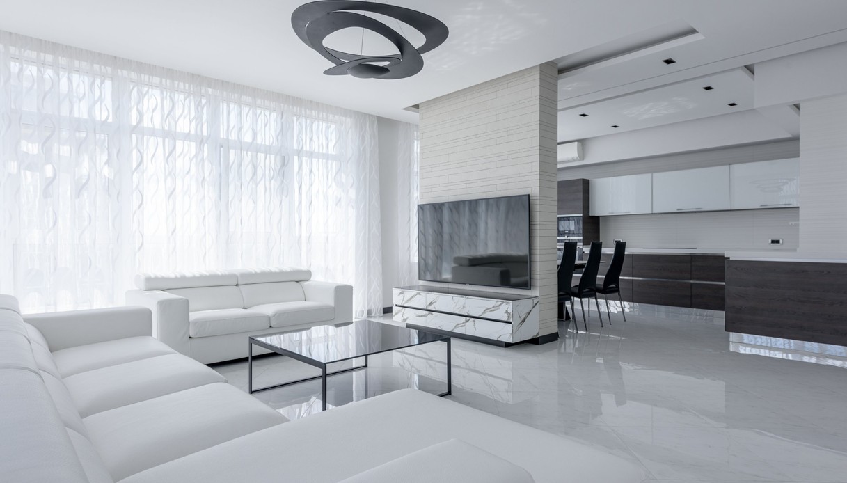 Soggiorno total white con la lampada a sospensione Pirce di Artemide, a spirale in metallo scuro, divani in pelle bianca, tavolino in vetro nero, pavimento lucido effetto marmo e cucina a vista sullo sfondo