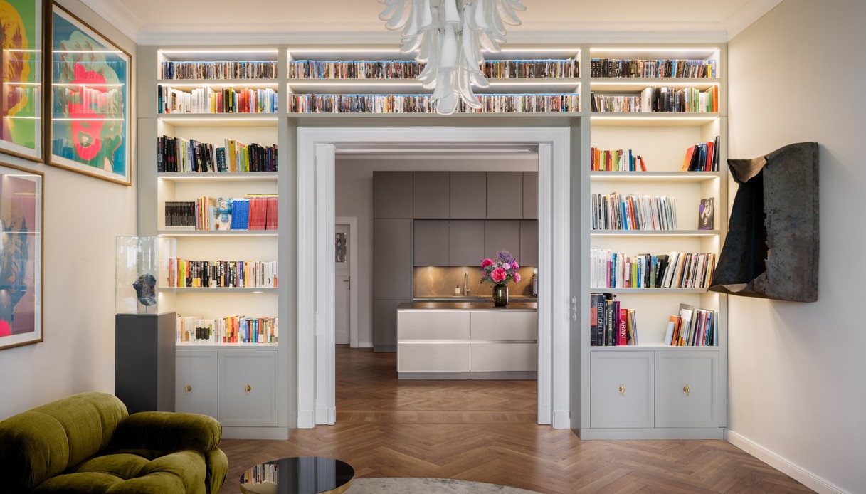Libreria illuminata a tutta parete attorno a un passaggio con cornice bianca, che lascia vedere la cucina con isola chiara e un vaso di fiori; a sinistra serigrafie colorate e una teca con un minerale, a destra un&rsquo;opera a parete scura