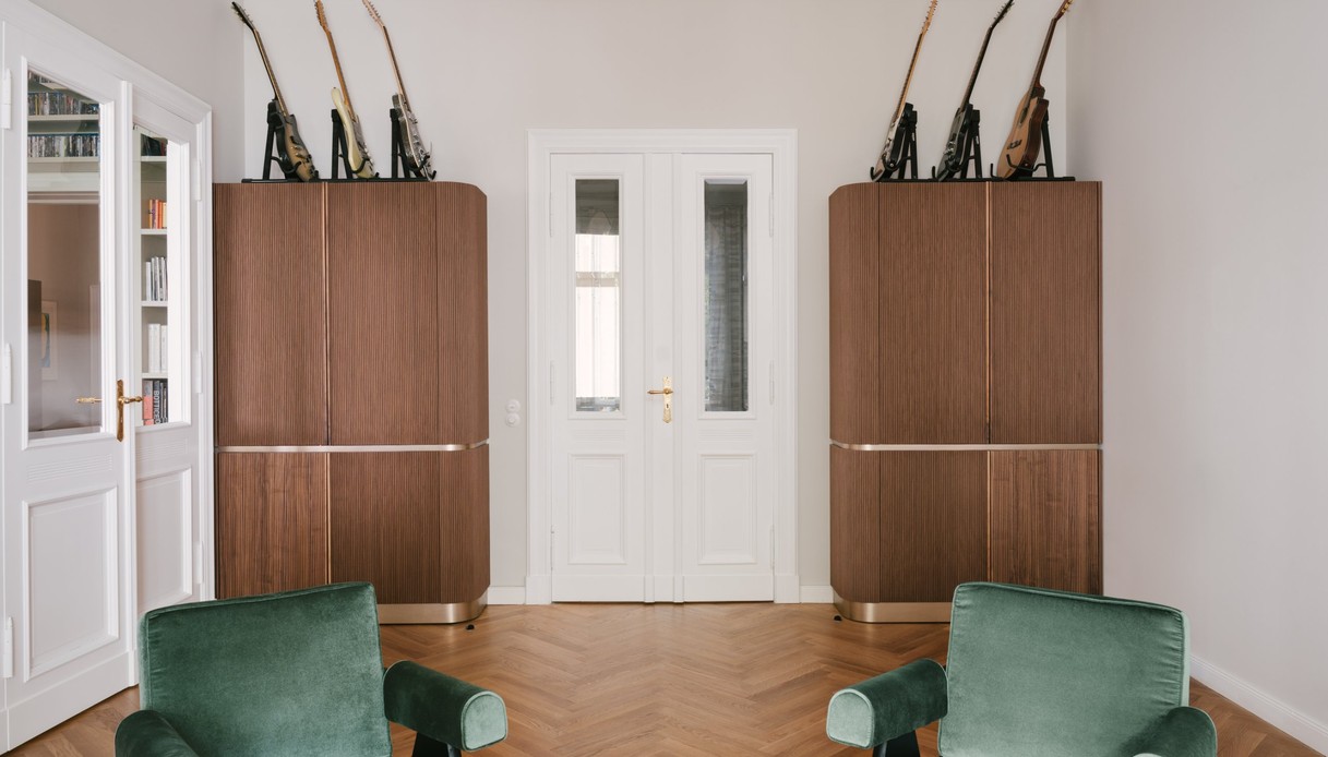 Stanza con due poltrone in velluto verde in primo piano, due mobili alti in legno scanalato con zoccolo dorato ai lati di una porta bianca a due ante, chitarre elettriche e acustiche esposte sopra i mobili e lampada a sospensione in fibra intrecciata