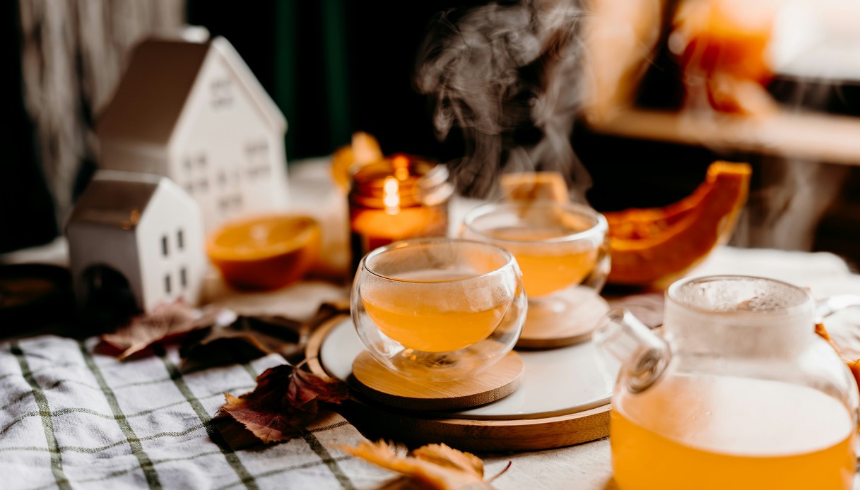 Tazze di vetro con tisana fumante su vassoio in legno, tovaglia chiara a quadretti verdi, foglie secche, candela accesa, zucca e casette in ceramica bianca