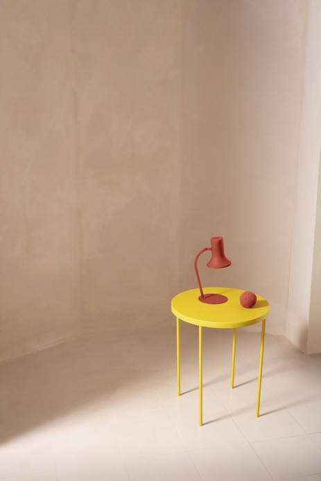 Tavolino rotondo giallo con lampada da tavolo color terracotta, in un ambiente con pareti e pavimento chiari dalla finitura continua