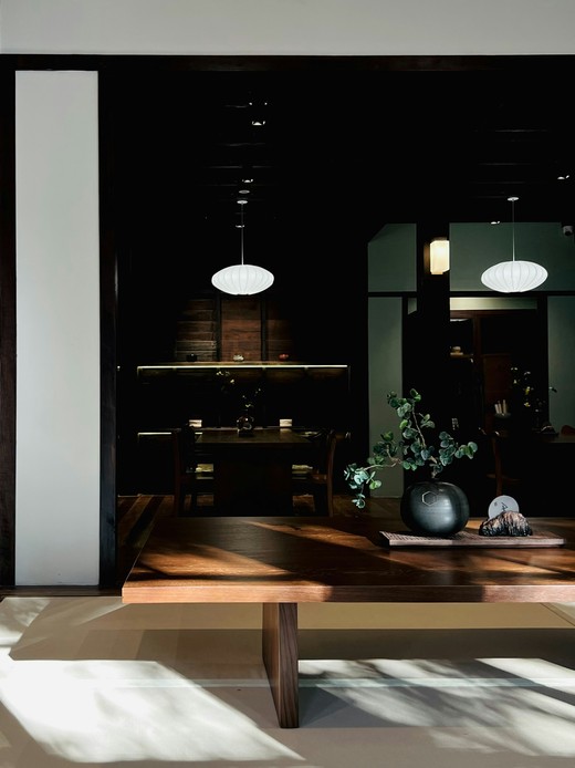 Tavolo basso in legno di noce illuminato da luce naturale radente, vaso scuro con ramo verde e lampade a sospensione bianche sullo sfondo