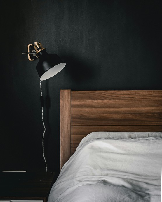 Testata del letto in legno con lenzuola bianche, parete dipinta di nero e applique nera con dettagli in ottone