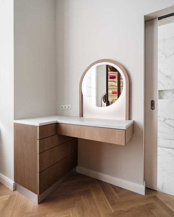 Toeletta angolare con cassettiera in legno scanalato, piano in marmo bianco venato e specchio tondo retroilluminato in una cornice ad arco di legno; a destra una porta scorrevole aperta su un bagno in marmo